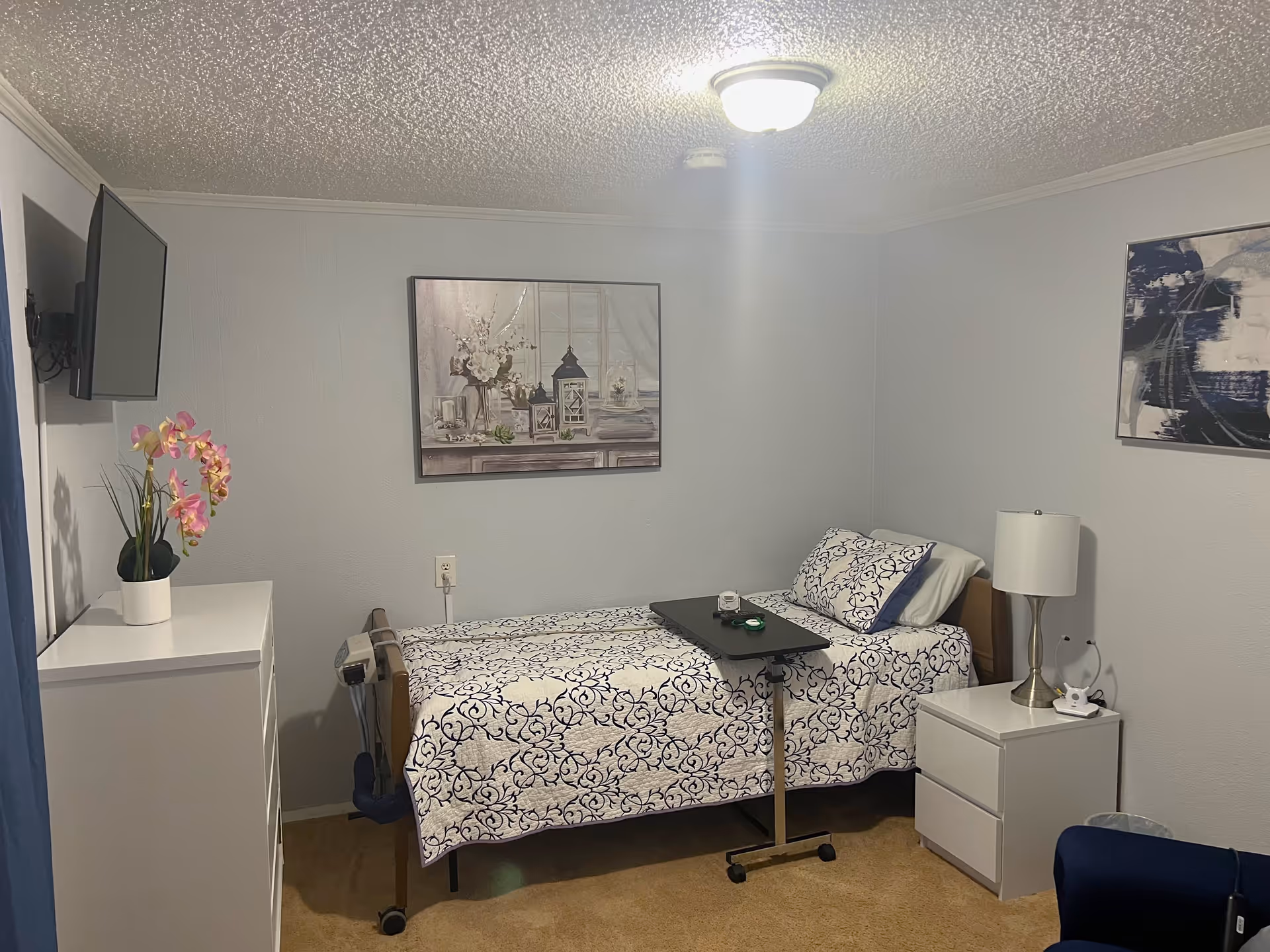 A small, neatly arranged bedroom with a single bed covered in a white and blue patterned quilt. The bed has a black overbed table with some items on it. To the right of the bed is a white nightstand with a lamp and a small device. On the left side of the room is a white dresser with a pink orchid plant on top and a wall-mounted flat-screen TV above it. The walls are light gray with two framed pictures hanging, one above the bed and one on the adjacent wall. The room has beige carpet and a ceiling light fixture.
