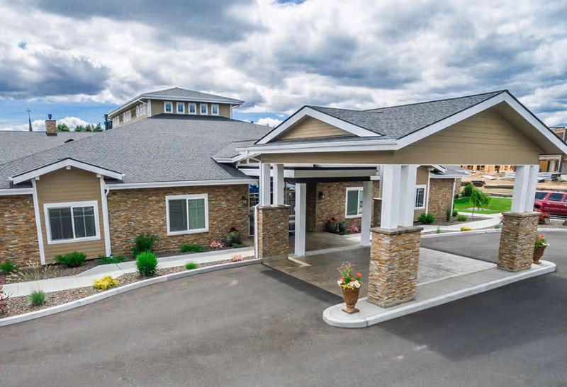 Exterior view of a senior living facility with a covered entrance supported by stone and white pillars. The building has beige siding with stone accents and multiple windows. There are landscaped areas with small bushes and flowers along the driveway under a cloudy sky.