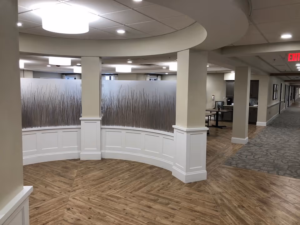 Interior view of a senior living facility hallway with wood-patterned flooring and carpet. There is a curved white half-wall with frosted glass panels featuring a grass design. The ceiling has recessed lighting and a large circular light fixture. The hallway extends into the distance with doors and framed pictures on the walls.