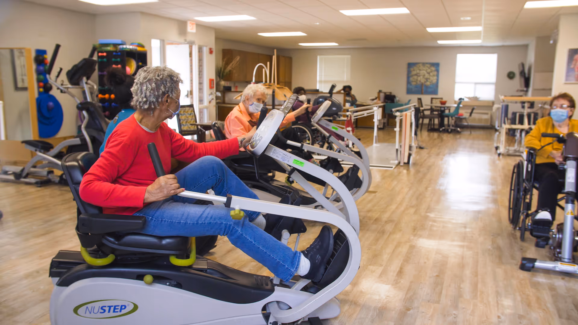 Several elderly individuals wearing masks are exercising on seated cardio machines in a spacious, well-lit fitness room with wooden floors and exercise equipment in the background.