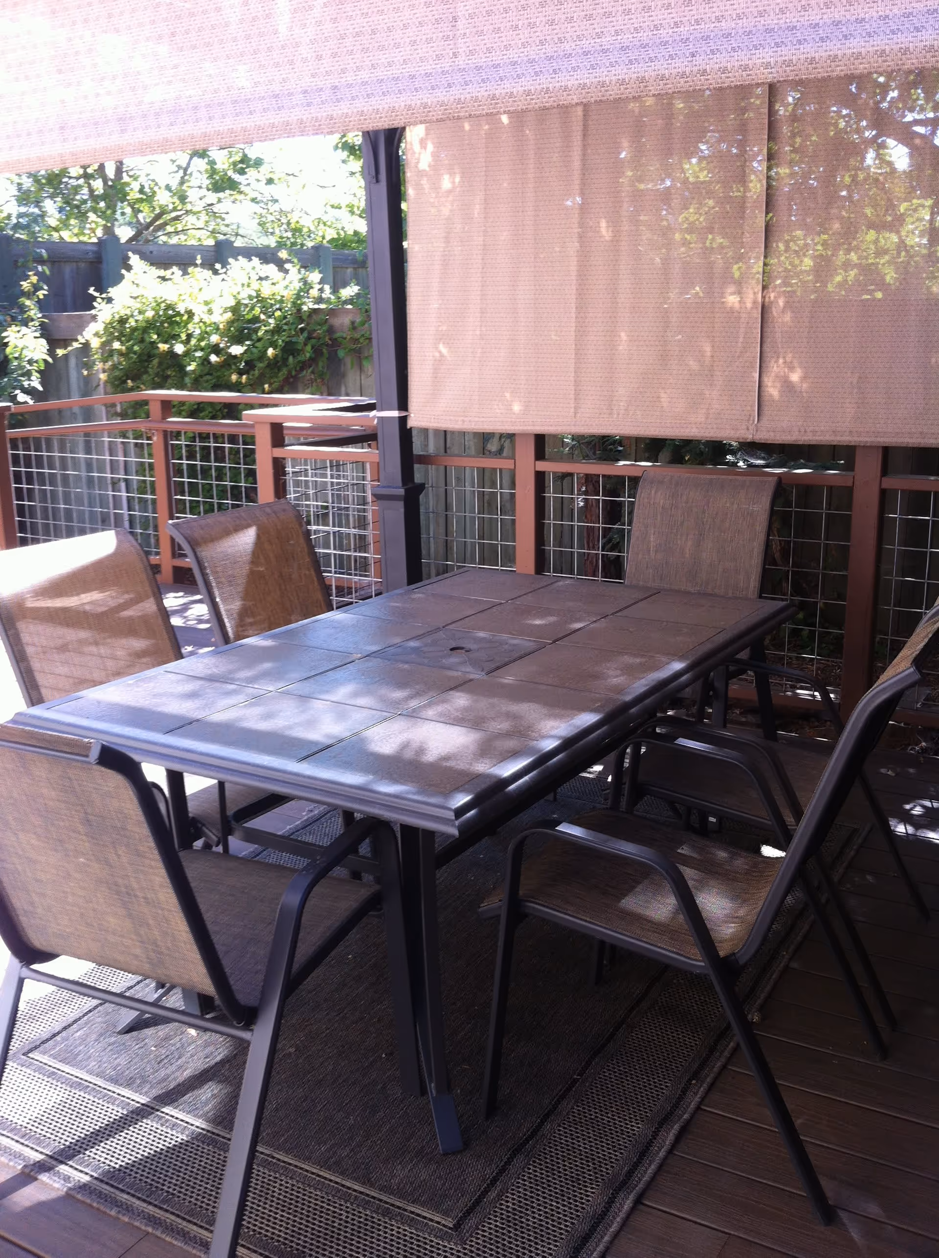 Outdoor covered patio with a rectangular dining table and six chairs on a wooden deck under a rolled shade.