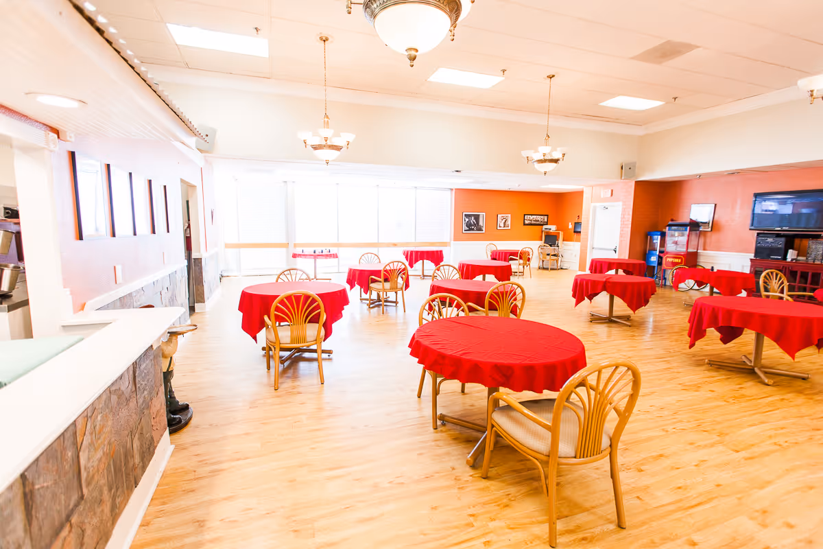 Bright dining/activity room with round tables covered in red tablecloths and wooden chairs on a wood floor.
