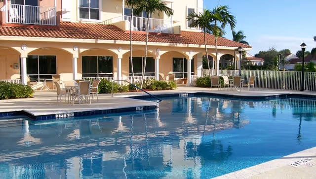 Outdoor swimming pool with clear blue water in front of a building with a red-tiled roof and balconies. Several tables and chairs are arranged on the pool deck, surrounded by palm trees and greenery under a clear blue sky.