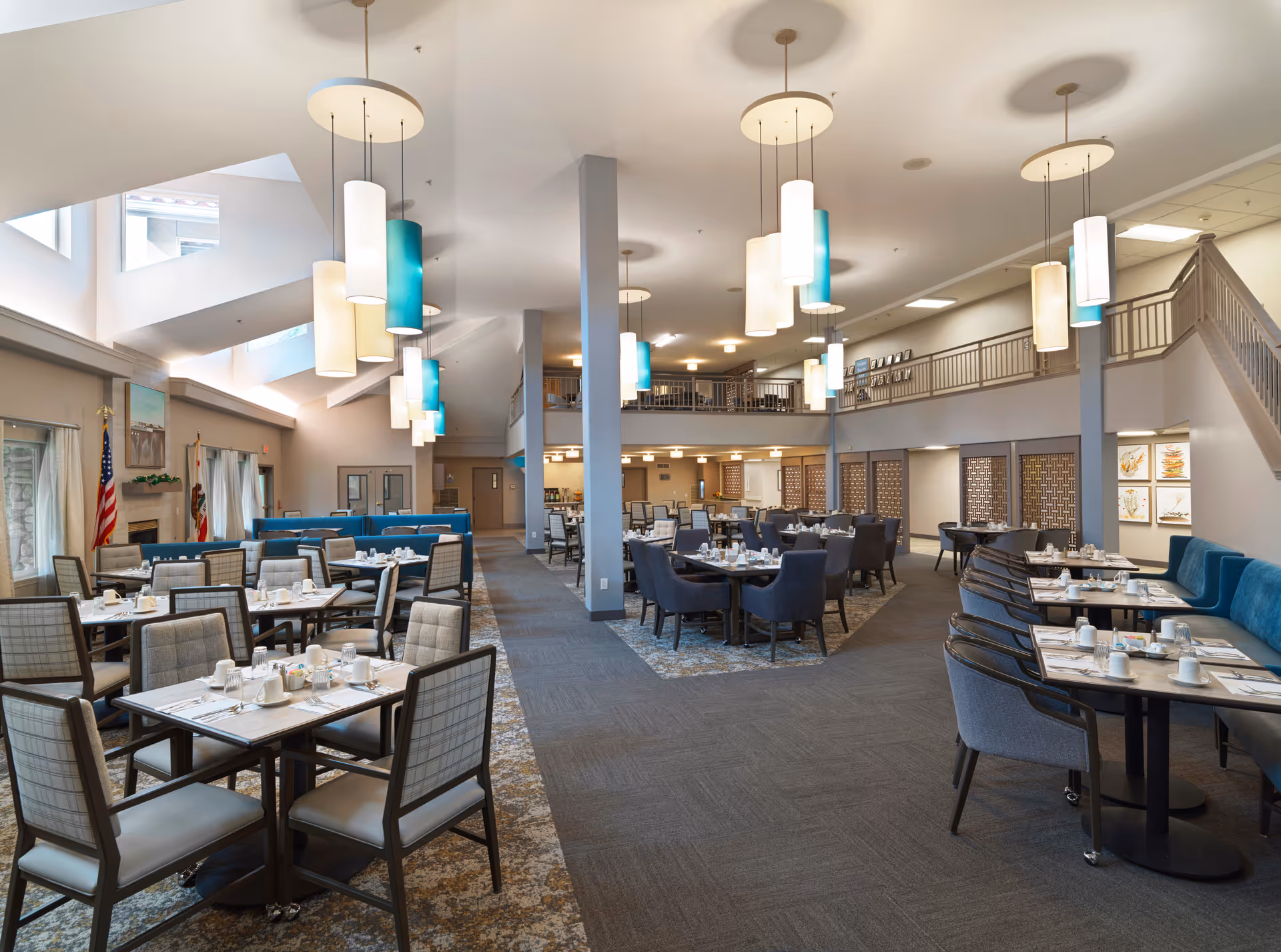 Spacious dining room in a senior living facility with multiple tables and chairs arranged neatly. The room features modern hanging light fixtures, large windows allowing natural light, and a high ceiling with skylights. Tables are set with cups, glasses, and utensils, ready for a meal service.