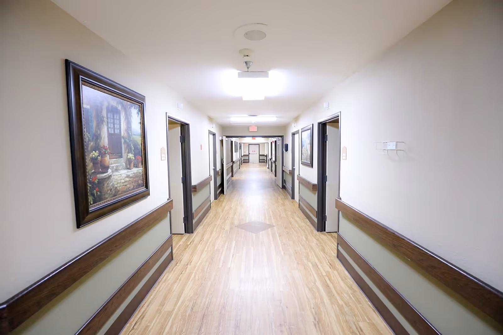 A long, well-lit hallway in a healthcare facility with light wood flooring, beige walls, handrails on both sides, and framed paintings hanging on the walls. Several doorways lead to rooms on either side of the corridor.