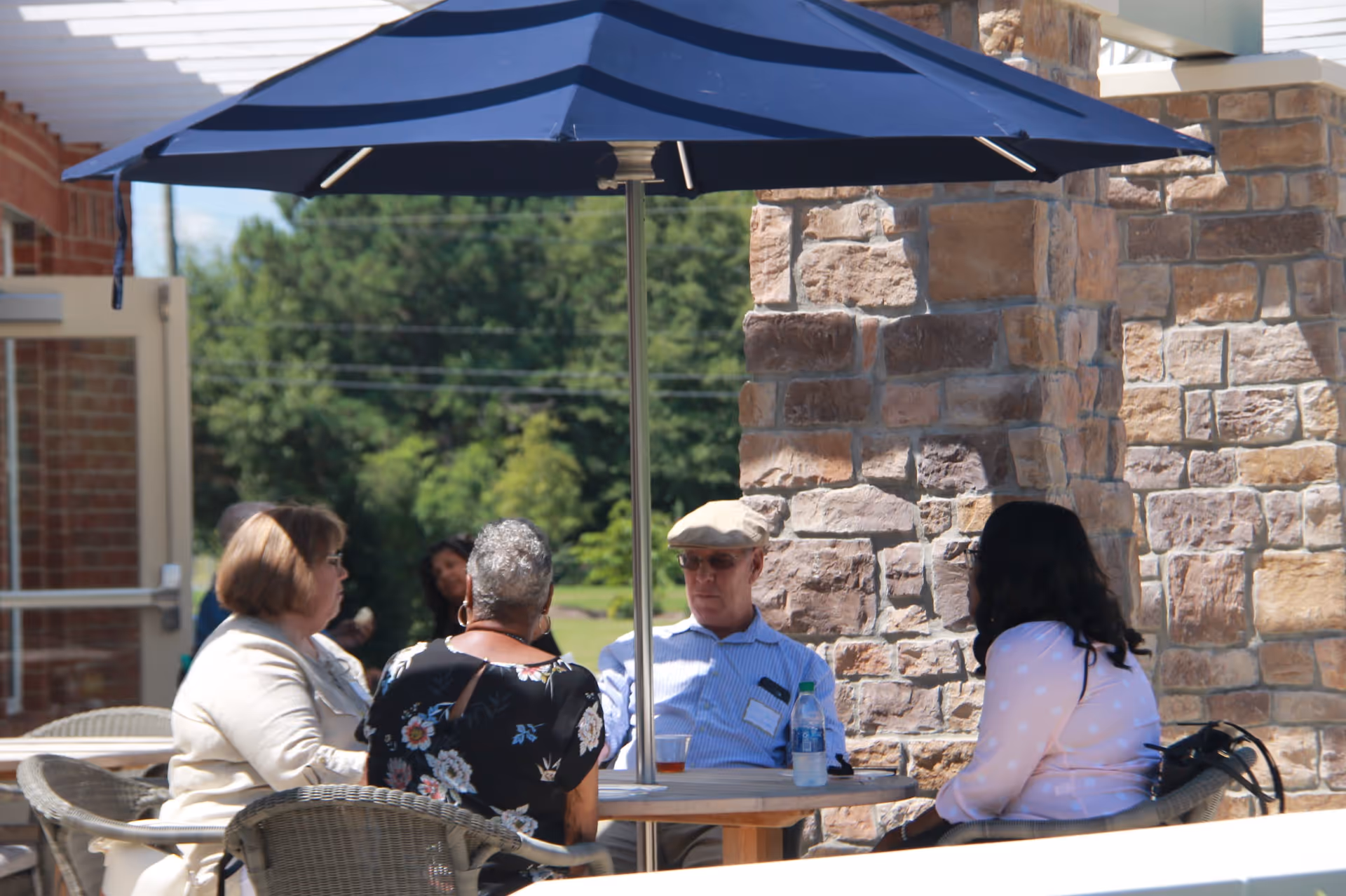 Four people sitting around a round outdoor table under a large blue umbrella, engaged in conversation. The setting is outside near a stone pillar and brick building, with greenery visible in the background.