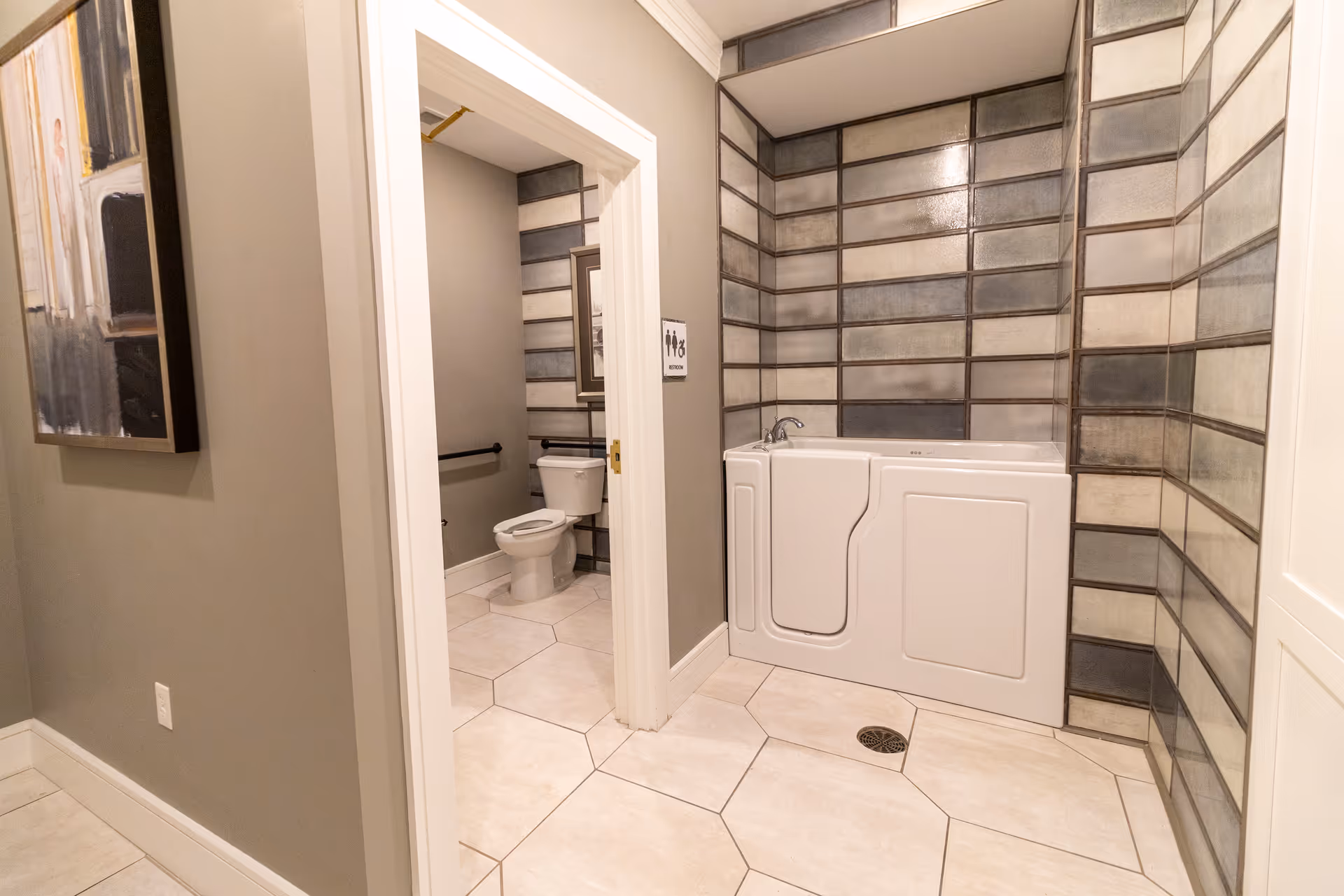 A bathroom area featuring a walk-in bathtub with tiled walls in shades of gray and beige. Adjacent to the bathtub is a separate toilet area with a white toilet, grab bars, and a framed picture on the wall. The floor is tiled with large hexagonal tiles, and the walls outside the bathroom are painted light gray with a piece of artwork hanging.
