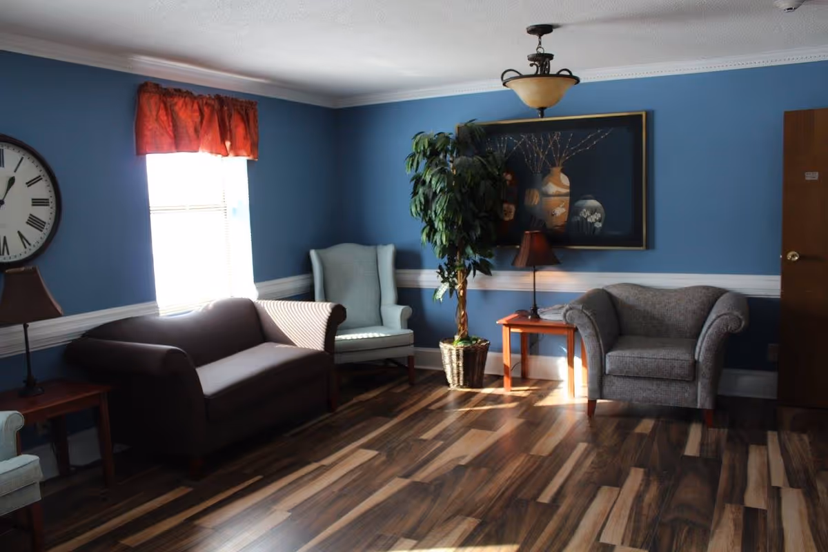 A cozy living room with blue walls and wood flooring. The room features a dark brown sofa, a light blue armchair, a gray armchair, two wooden side tables with lamps, a large wall clock, a potted plant, and a framed artwork on the wall. Sunlight streams in through a window with a red valance.