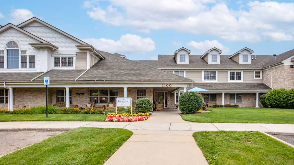Front exterior view of Brookdale Portage facility showing a two-story building with a covered entrance, manicured lawn, flower beds, and a clear blue sky with some clouds.