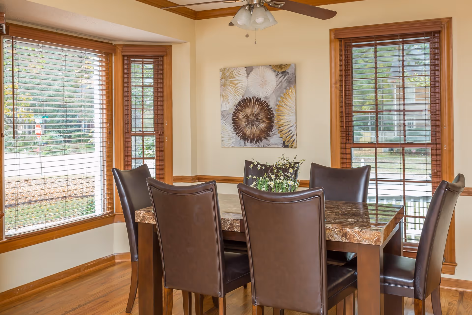 A dining room with a rectangular marble-top table surrounded by six dark brown leather chairs. The room has large windows with wooden blinds, light beige walls, and a ceiling fan with lights. A decorative painting featuring abstract floral designs hangs on the wall.