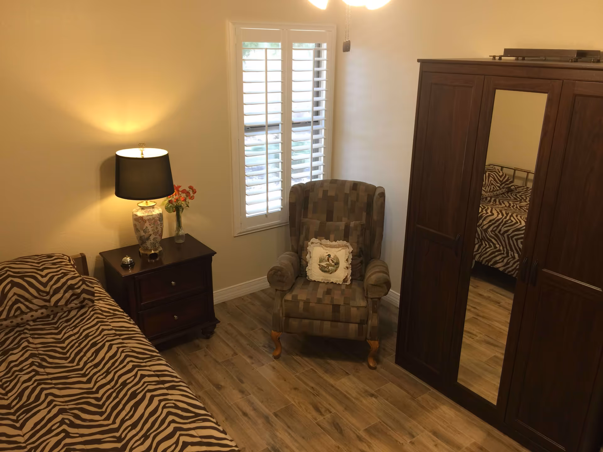 A cozy bedroom with a single bed covered in zebra print bedding, a wooden nightstand with a decorative lamp and a small vase of flowers, a patterned armchair with a decorative pillow, a window with white shutters, and a large wooden wardrobe with a mirrored door reflecting part of the bed.