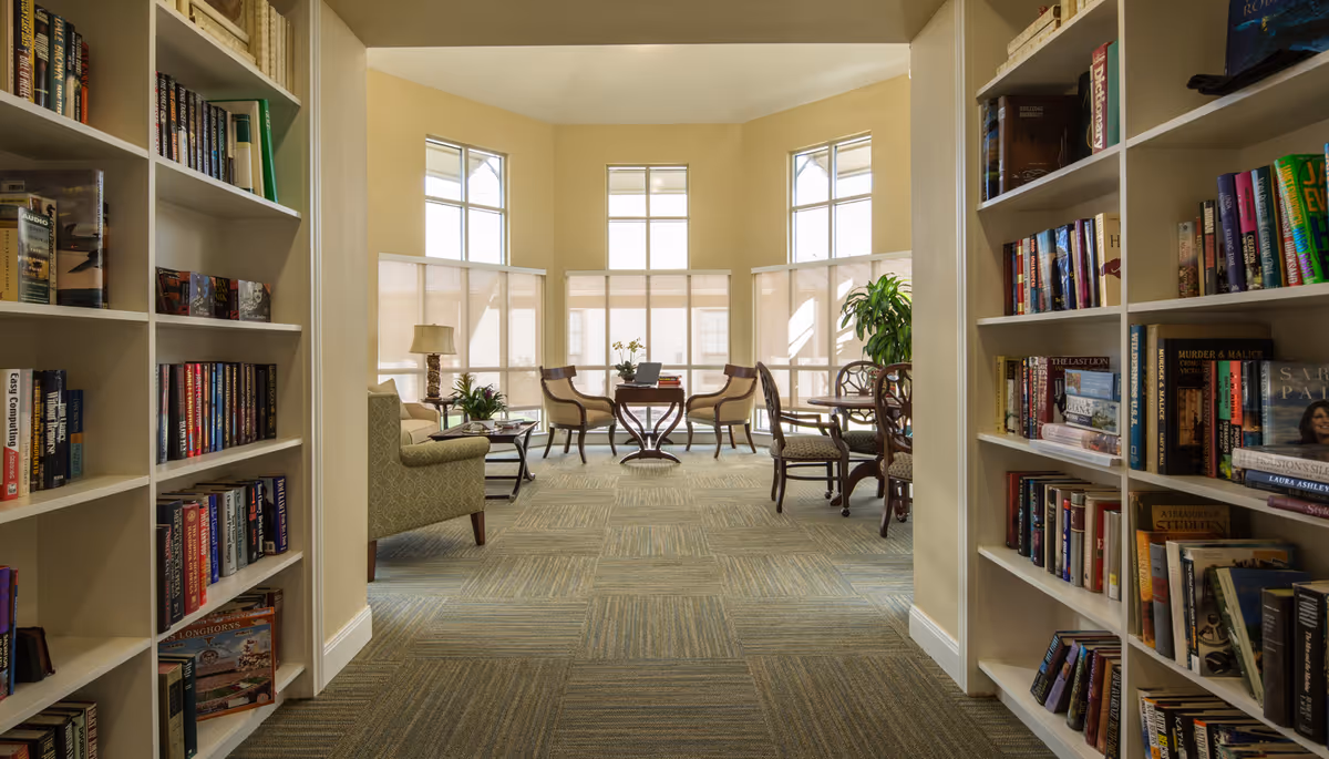 A bright and inviting library or reading room with bookshelves filled with books on both sides of the entrance. The room has large windows with blinds partially drawn, allowing natural light to fill the space. There are several chairs and small tables arranged for reading or conversation, along with a few plants and a lamp adding to the cozy atmosphere.