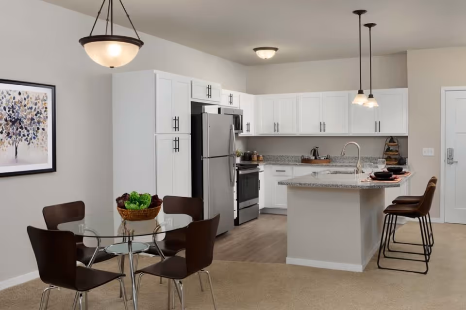 Open-concept kitchen and dining area with white cabinets, a granite island with bar stools, stainless appliances, and a round glass dining table.