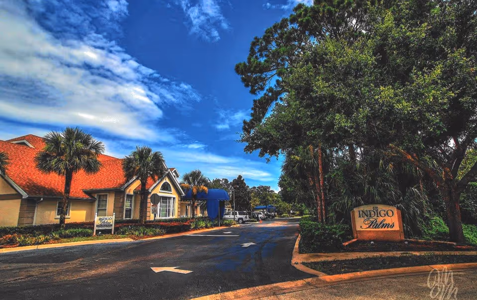 Exterior view of Indigo Palms facility with a driveway, palm trees, and a building with a red roof under a partly cloudy blue sky.