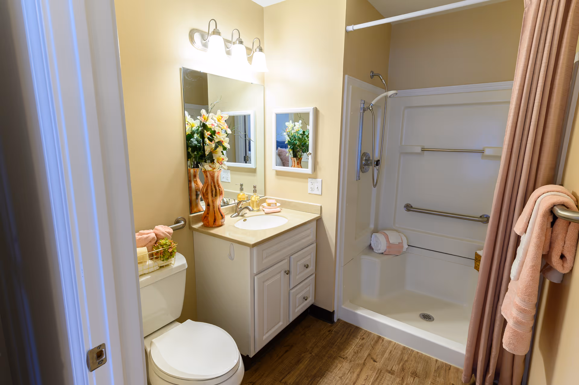 Small, well-lit bathroom featuring a vanity with mirror and flowers, a toilet, and a walk-in shower with grab bars and towels.