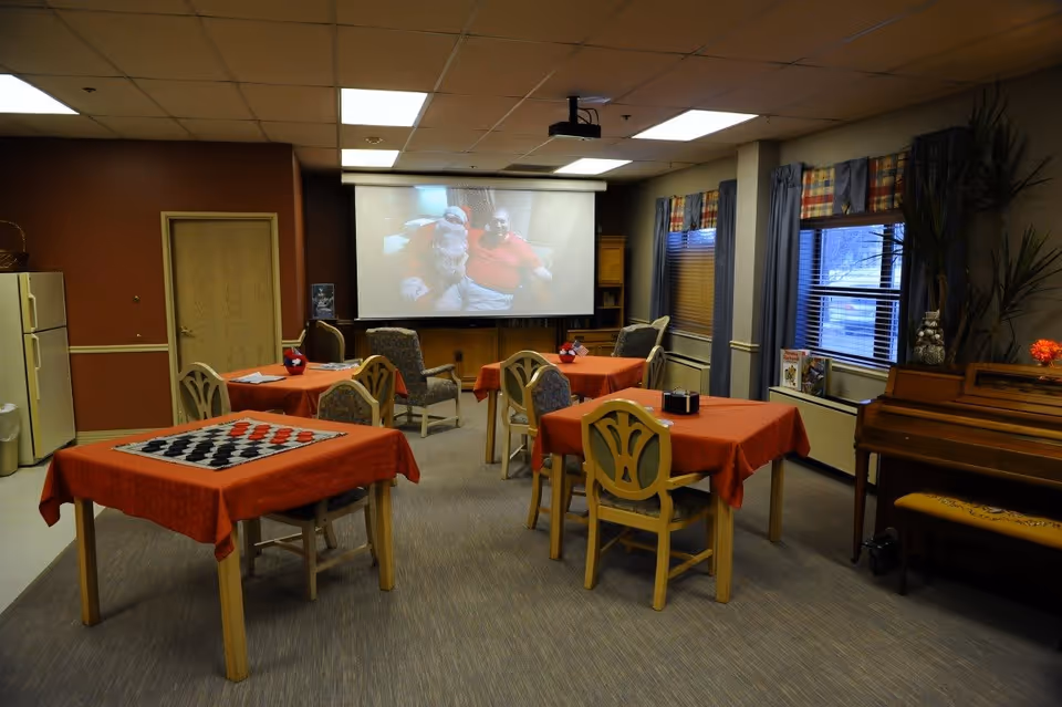 A common room with several tables covered in orange tablecloths, each surrounded by chairs. One table has a checkers board on it. There is a piano with a bench on the right side near windows with blue curtains. A projector screen at the far end displays an image of two people sitting together. The room has a beige carpet and a ceiling with fluorescent lights.
