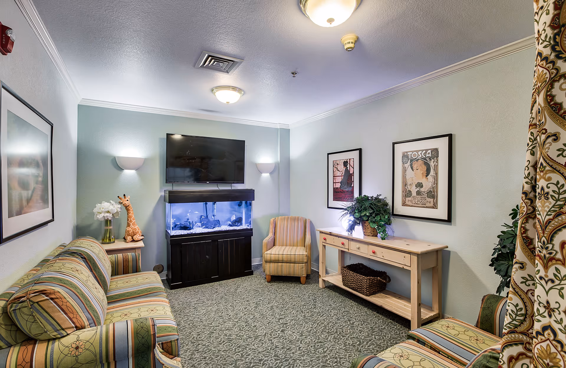 A cozy living room with a striped sofa and armchair, a large fish tank beneath a wall-mounted flat screen TV, framed artwork on the walls, a wooden console table with a basket and plants, and patterned curtains on the right side.