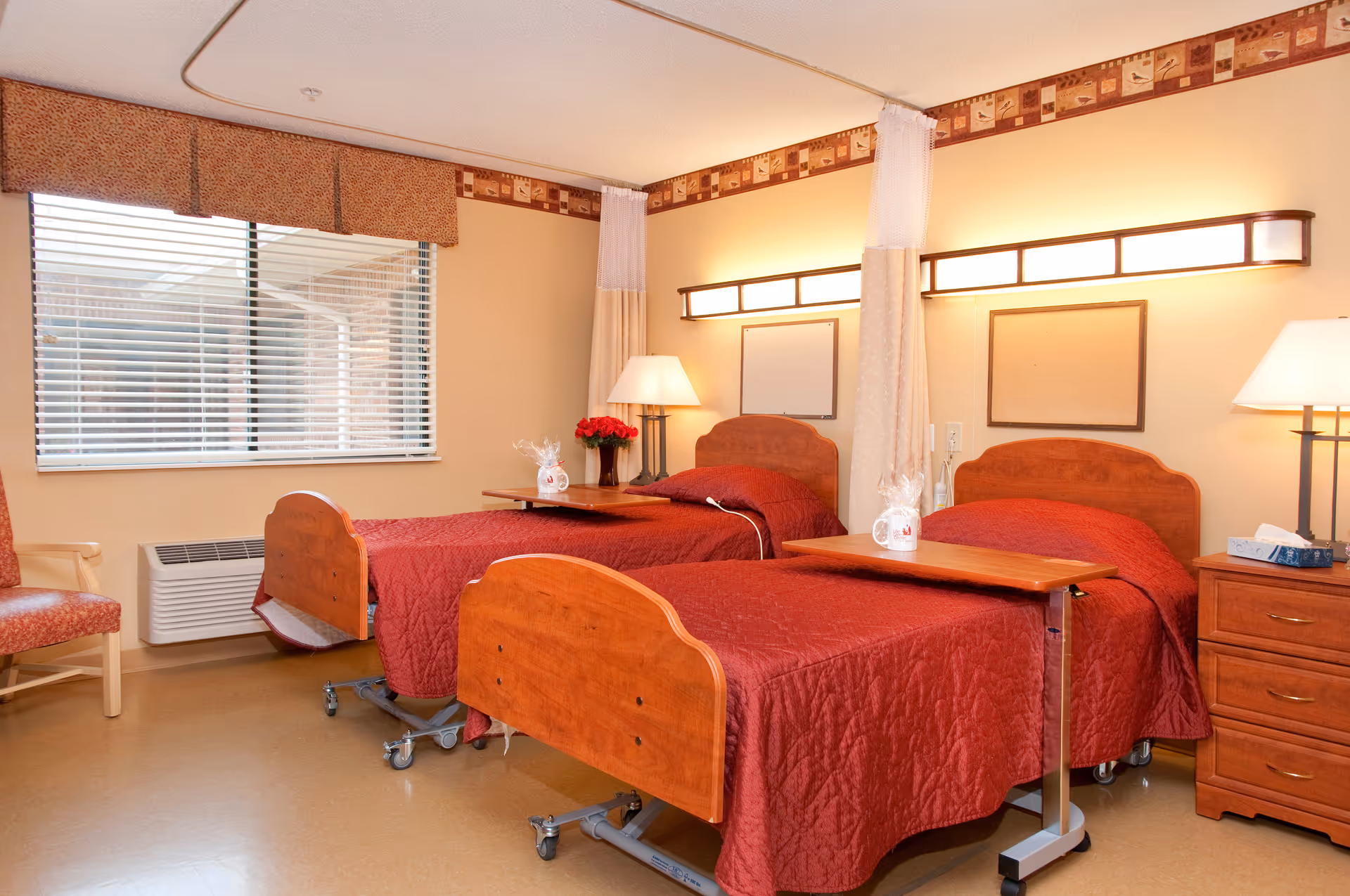 A bright senior living bedroom with two hospital-style beds covered in red quilts, bedside tables, lamps, and a window.