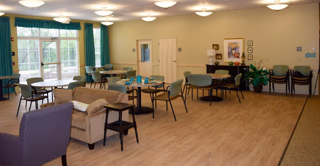A spacious, well-lit common area with multiple round tables and chairs arranged for dining or socializing. There are large windows with teal curtains letting in natural light, a beige couch and purple armchair, and a sideboard with decorative items and a framed picture on the wall. The floor is wood, and the walls are painted a light beige color.