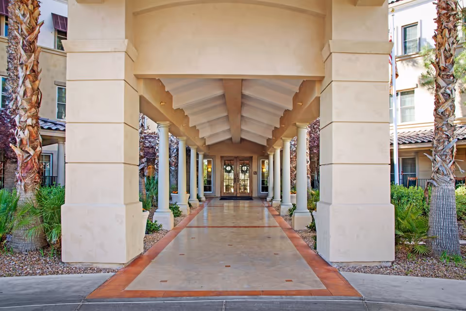 Covered walkway entrance to a building with beige columns and a tiled floor, flanked by palm trees and greenery, leading to double glass doors with wreaths.
