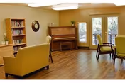 A cozy living room area with wooden flooring, featuring a piano against the wall, a bookshelf filled with books, a round mirror, and several chairs and armchairs arranged for seating. Large windows and glass doors allow natural light to enter the room.