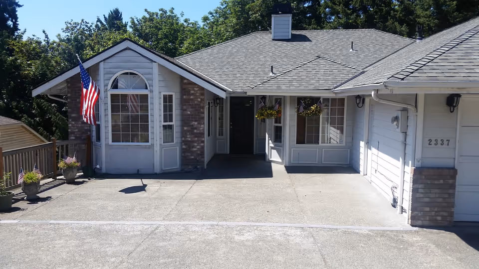 Front exterior view of a single-story senior living facility building with a gray shingled roof, white siding, and brick accents. The entrance area has hanging flower pots with small American flags, and a larger American flag is mounted near a window. The building number 2337 is visible on the right side near the garage door. The surrounding area includes trees and a wooden fence with potted plants.