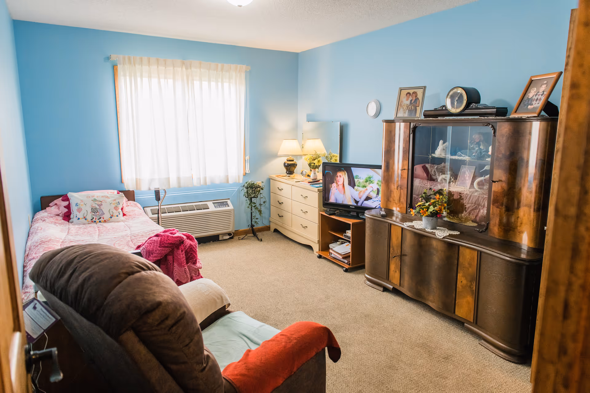 Sunny pastel-blue furnished bedroom with a single bed, recliner, dresser, TV, and a wooden display cabinet.