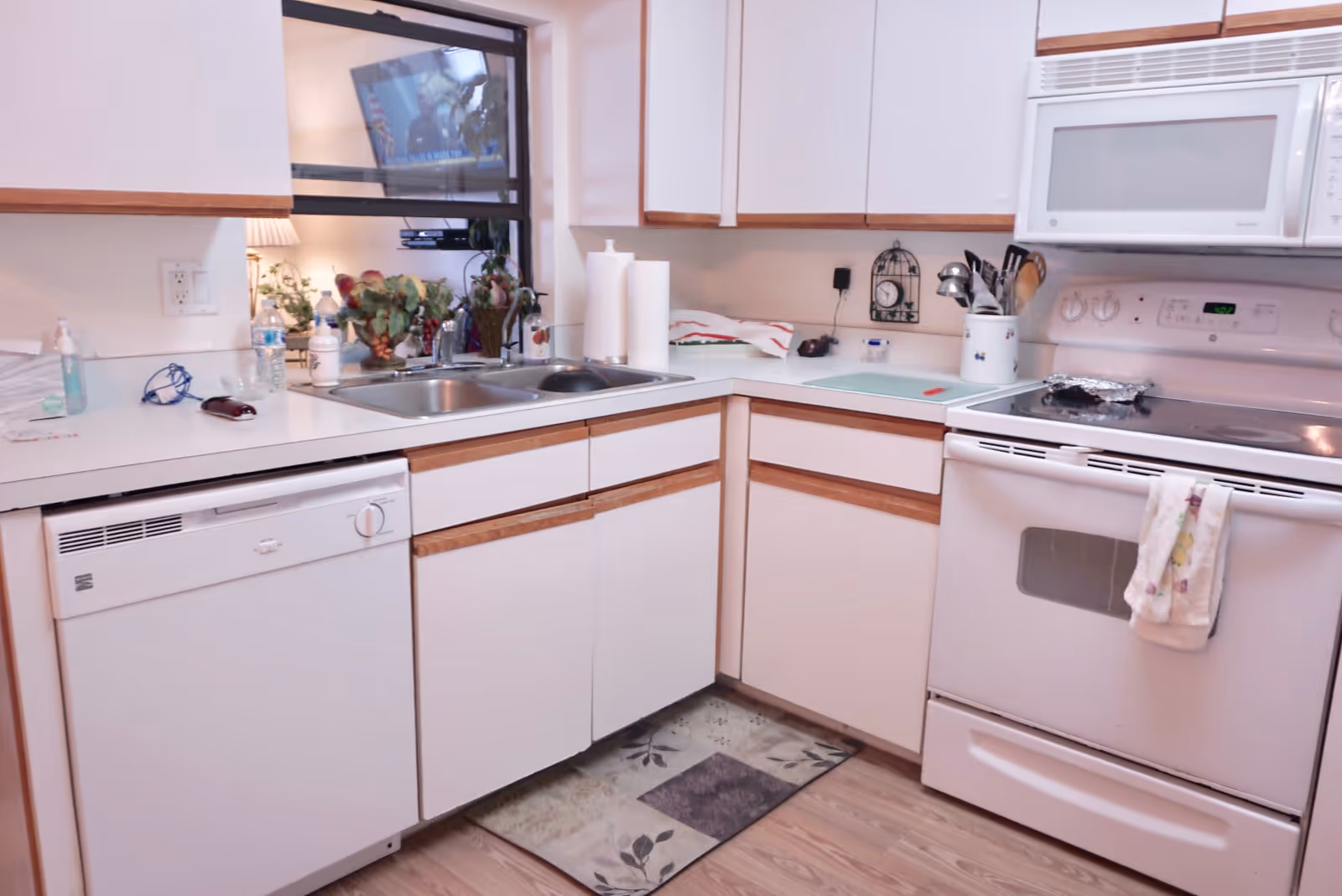 A clean kitchen with white cabinets and wooden trim, featuring a dishwasher, double sink, electric stove with oven, microwave, and various kitchen utensils. There is a window above the sink showing a TV and some plants in the adjacent room.