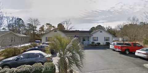 Parking lot with several parked cars in front of a single-story residential building surrounded by trees and bushes under a cloudy sky.