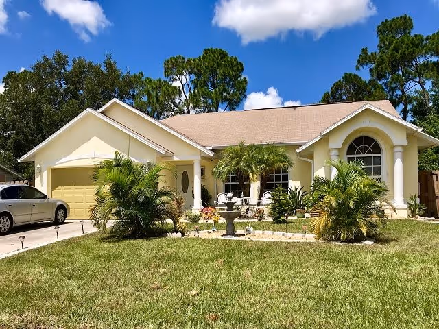 Front exterior view of a single-story residential building with a beige facade, a two-car garage, a driveway with a parked car, a well-maintained lawn, palm trees, and a decorative fountain in the garden under a blue sky with some clouds.