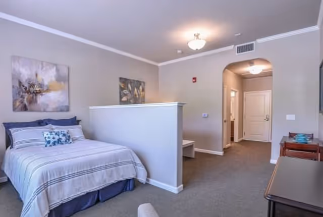 A spacious bedroom in a senior living facility featuring a neatly made bed with striped bedding and decorative pillows. The room has neutral-colored walls with two paintings hanging above the bed. A half-wall partially separates the sleeping area from the rest of the room, which includes a small bench, a desk with a chair, and a hallway leading to a white door. The room is carpeted and well-lit with ceiling lights.