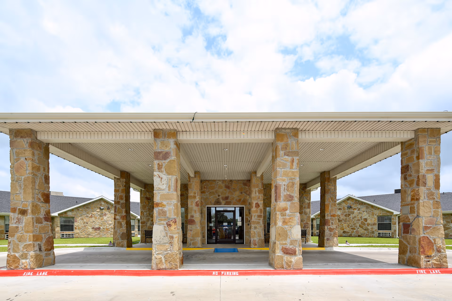 Front entrance of Windsor Nursing and Rehabilitation Center of Corpus Christi featuring a covered drop-off area supported by stone pillars, with a clear sky in the background.