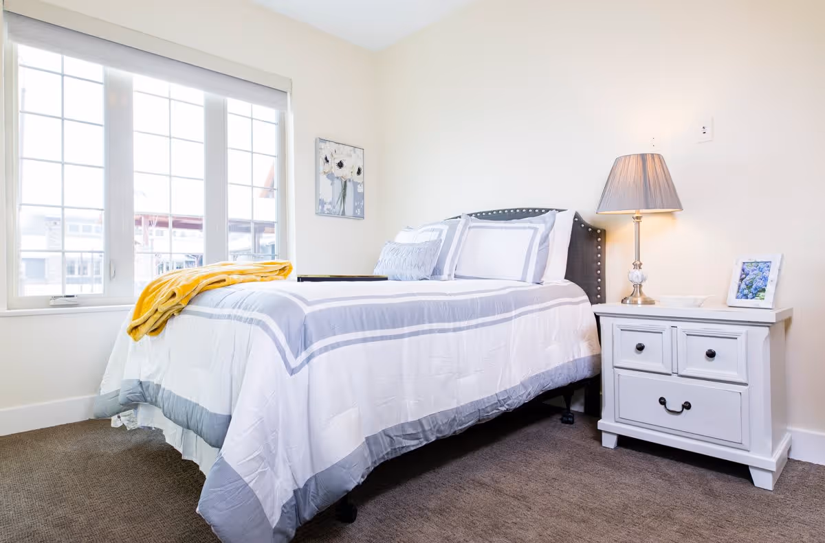 A bright bedroom with a neatly made bed featuring white and gray bedding, a yellow blanket folded at the foot of the bed, a white nightstand with drawers, a table lamp, and a framed picture. There is a large window with multiple panes letting in natural light and a floral painting on the wall.