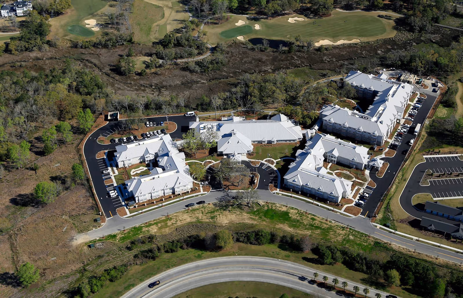 Aerial view of Wellmore of Daniel Island senior living facility showing multiple white-roofed buildings arranged around a circular driveway with parking lots, surrounded by greenery and a golf course in the background.