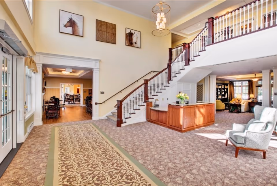 Spacious and well-lit senior living facility lobby with a patterned carpet, a wooden reception desk, a staircase with wooden banisters, and comfortable seating areas. The walls are decorated with framed horse artwork, and there is a chandelier hanging from the ceiling. The lobby opens into a dining area with tables and chairs.