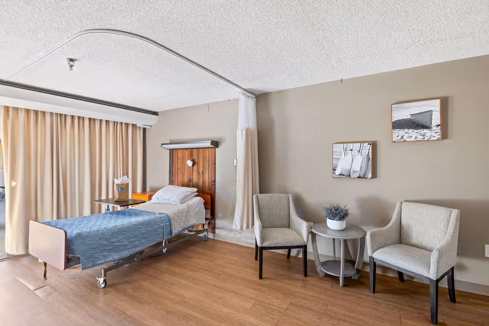 A senior care facility room with a single hospital-style bed covered with a blue blanket and white pillow. Next to the bed is a small wooden nightstand and a table with a paper bag on it. Two beige upholstered chairs flank a small round table with a potted plant. The walls are beige with two framed black and white photos of boats and oars. A curtain hangs from a ceiling track, and large beige curtains cover a window or sliding door.