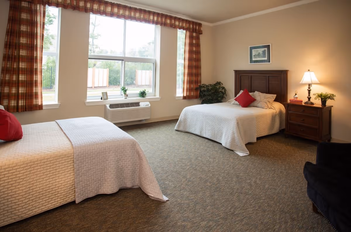 A spacious bedroom with two beds covered in white quilts, each with red accent pillows. The room has large windows with plaid curtains, a wooden nightstand with a lamp, books, and a plant, and a framed picture on the wall. There is also a black armchair and a green plant in the corner.