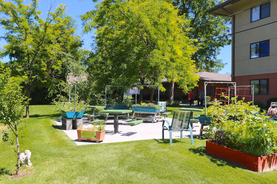 A sunny outdoor garden area at a senior living facility with green grass, trees, raised garden beds with plants, picnic tables, and chairs. A building is visible on the right side and two people are walking in the background.
