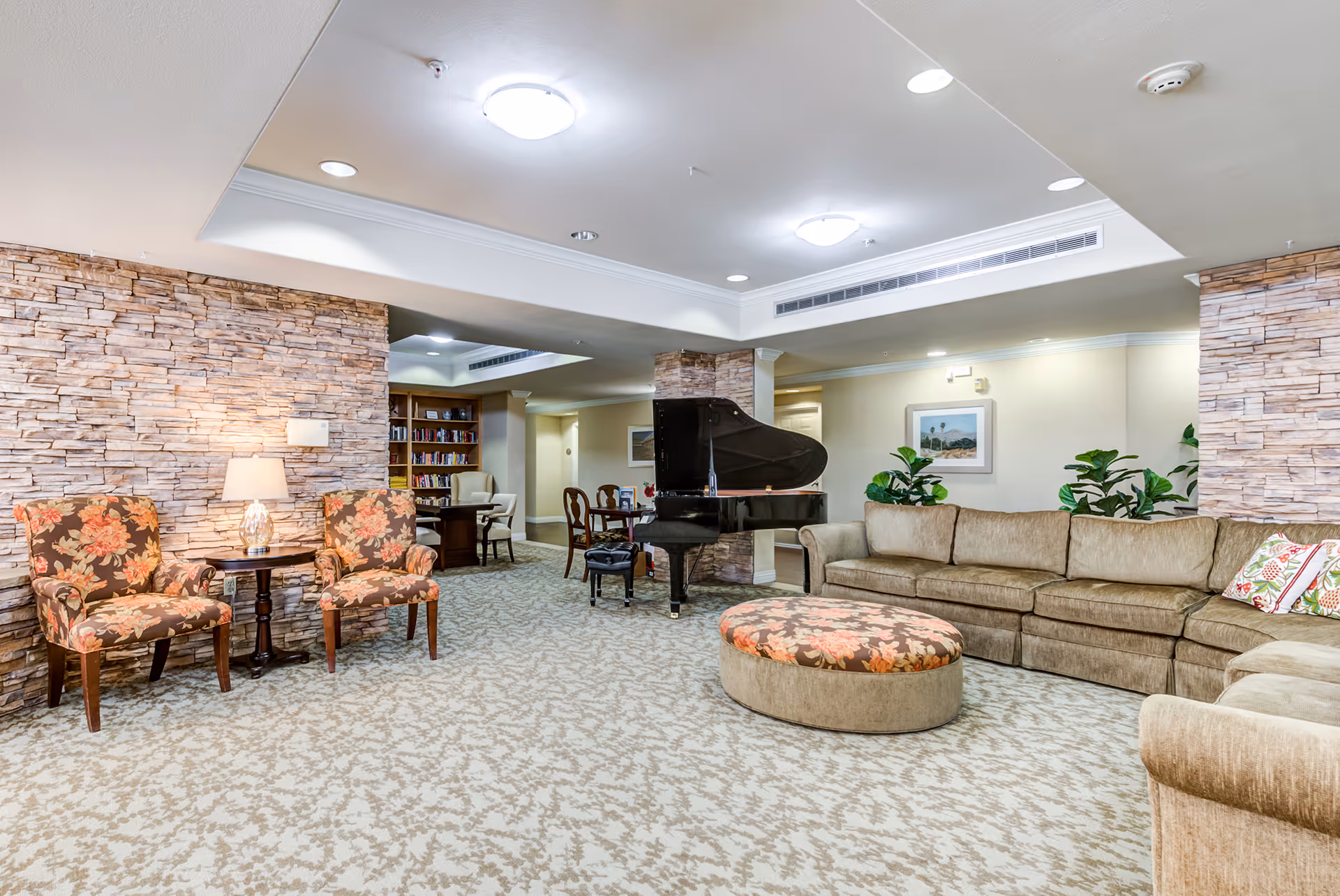 A spacious living room area in a senior living facility with a large beige sectional sofa, a round floral ottoman, two floral upholstered chairs with a small table and lamp between them, a black grand piano, and a bookshelf in the background. The walls have a stone accent and there are several plants and framed artwork on the walls.