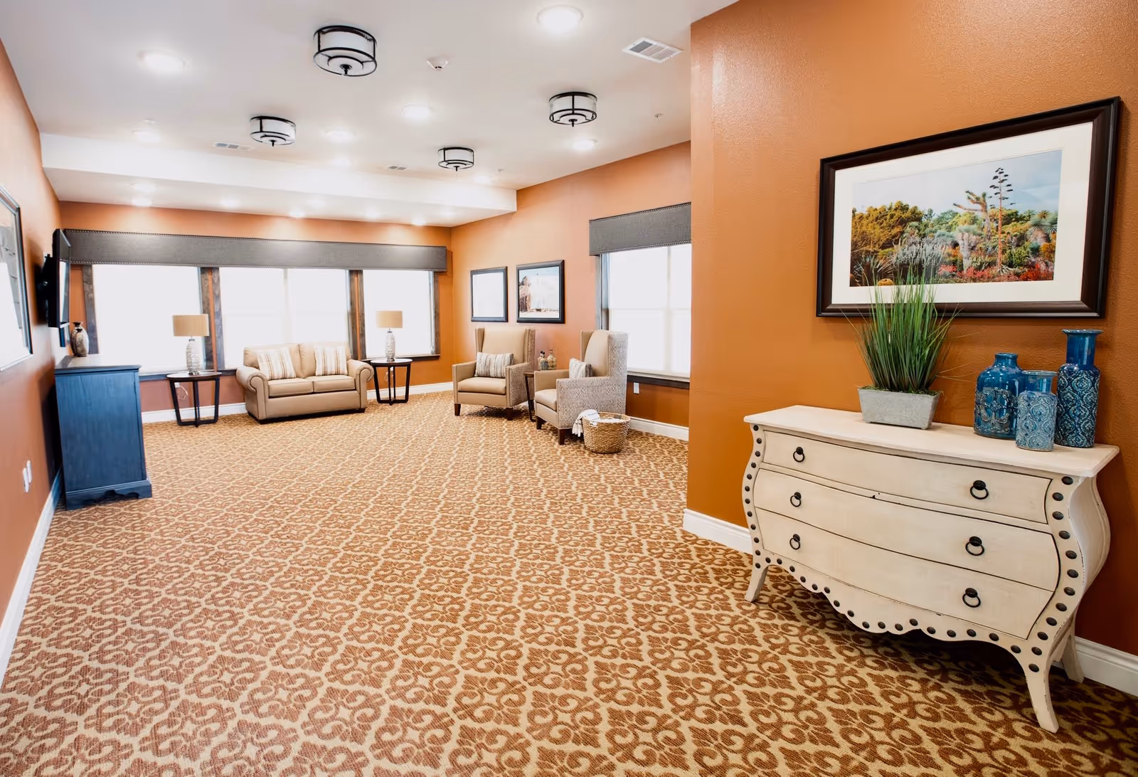 A spacious, well-lit common area with patterned carpet, orange walls, and multiple seating options including a beige sofa and two armchairs. There are side tables with lamps near the windows, a TV mounted on the wall, and a decorative chest with blue vases and a plant. Framed artwork hangs on the walls.
