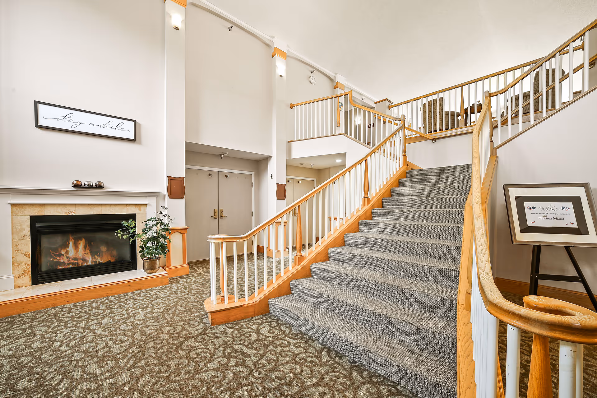 Interior view of a senior living facility lobby featuring a carpeted staircase with wooden handrails leading to an upper level. There is a lit fireplace with a plant beside it on the left, a framed sign above the fireplace that reads 'stay awhile', and a welcome sign on an easel to the right of the staircase. The area has patterned carpet and neutral-colored walls.
