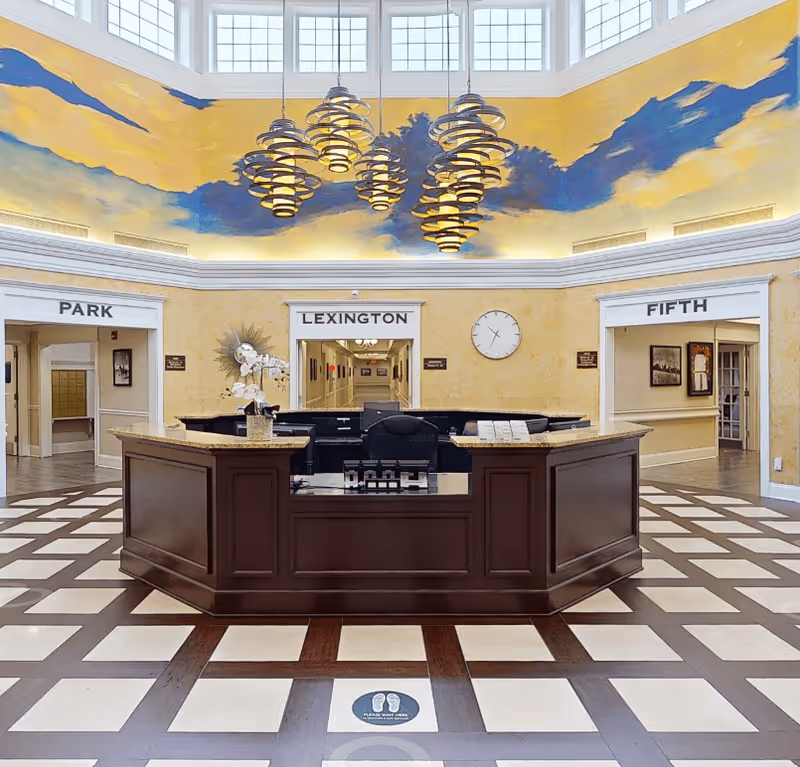 Reception area with a large dark wood desk in the center, surrounded by a checkered floor. Above the desk are modern circular pendant lights hanging from a ceiling with a mural of a blue sky and clouds. Three doorways labeled Park, Lexington, and Fifth lead to different hallways. A clock is mounted on the wall above the Lexington doorway.