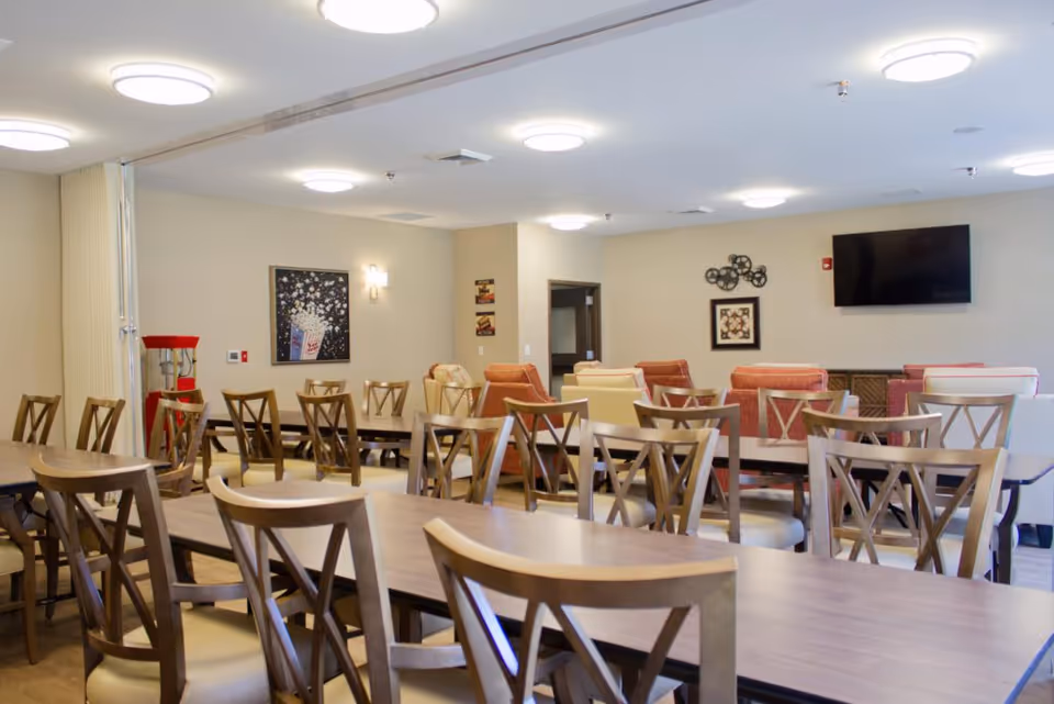 A spacious dining room with multiple wooden tables and chairs arranged neatly. The room features beige walls with framed artwork, a popcorn machine in the corner, and a flat-screen TV mounted on the wall. Comfortable armchairs are placed along the back wall, creating a cozy seating area.