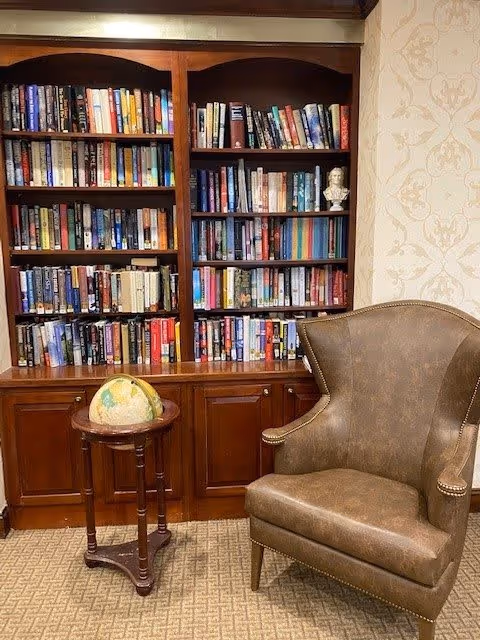 A cozy reading nook featuring a tall wooden bookshelf filled with books, a small wooden side table with a globe on top, and a brown leather armchair with nailhead trim, set against a wallpapered wall with a subtle floral pattern.