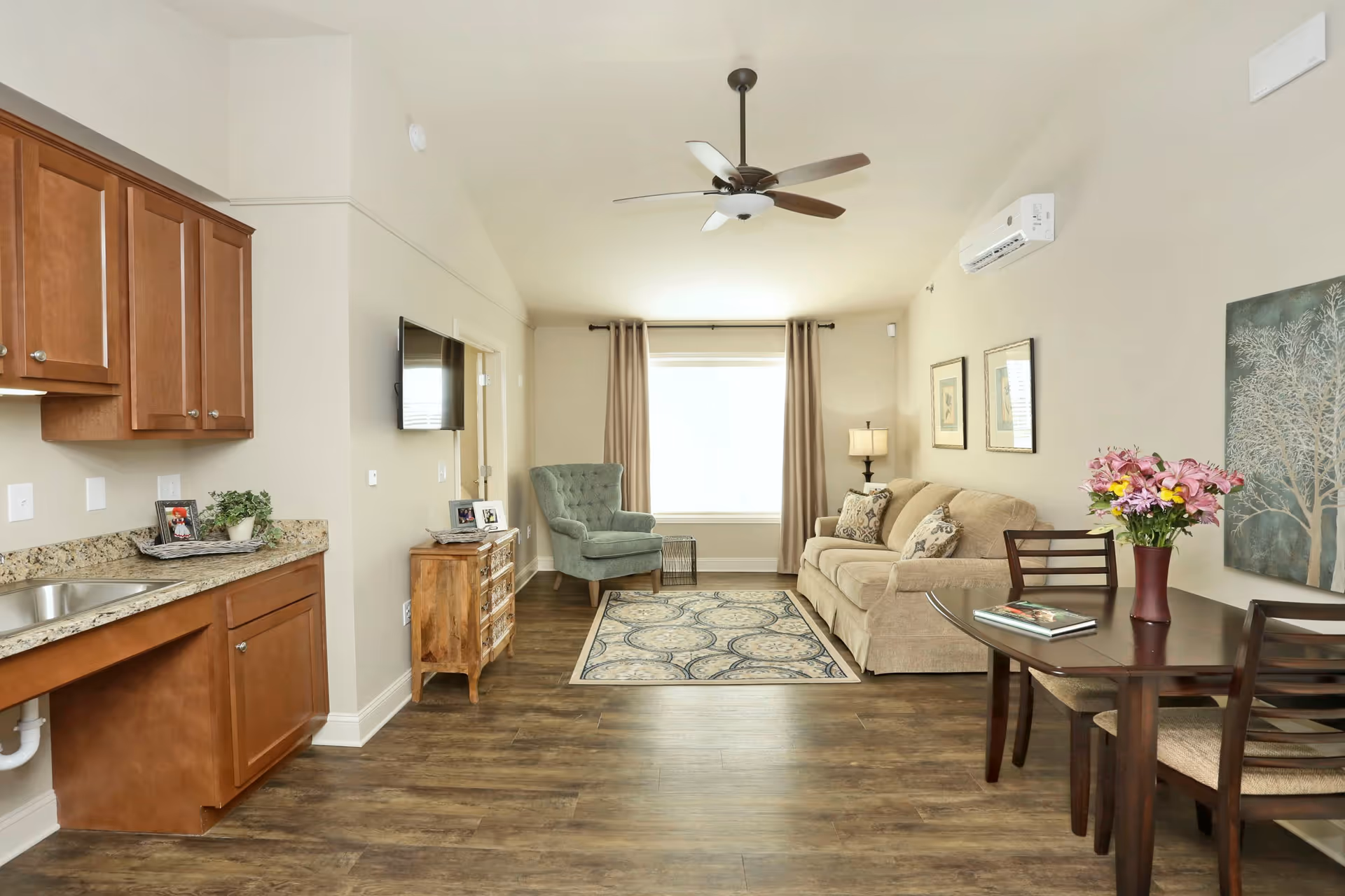 A bright and cozy living area in an assisted living facility featuring a beige sofa with patterned cushions, a green upholstered armchair, a wooden side table with framed photos, a wall-mounted flat screen TV, a ceiling fan, and a window with beige curtains. The room also includes a small dining table with two chairs and a vase of flowers, wooden cabinets with a granite countertop and a sink, and framed artwork on the walls.