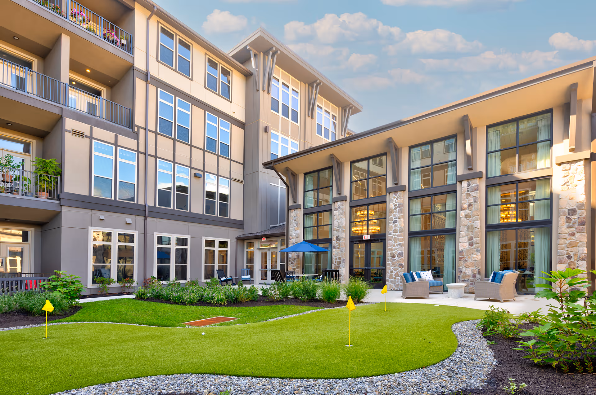 Outdoor courtyard area of a senior living facility with a putting green, patio seating with chairs and tables, surrounded by a multi-story building with large windows and balconies. The sky is clear with some clouds.