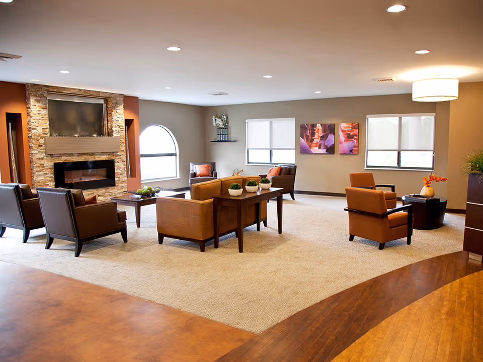 A spacious and well-lit living room area with multiple brown armchairs and sofas arranged around a central coffee table and side table. The room features a stone fireplace with a modern electric insert, large windows with blinds, and warm-toned walls. Decorative plants and artwork add to the cozy ambiance.