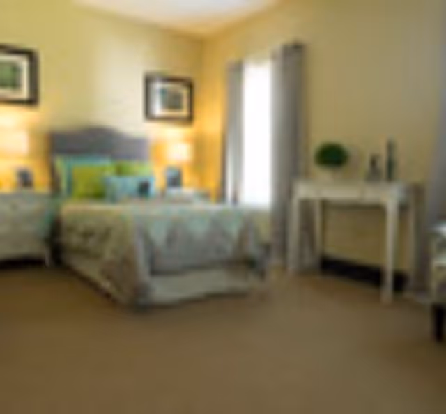 A cozy bedroom with a bed featuring a patterned bedspread and multiple pillows. There are two nightstands with lamps on either side of the bed, framed pictures on the wall above the headboard, a window with curtains, and a small white desk with decorative items on it.