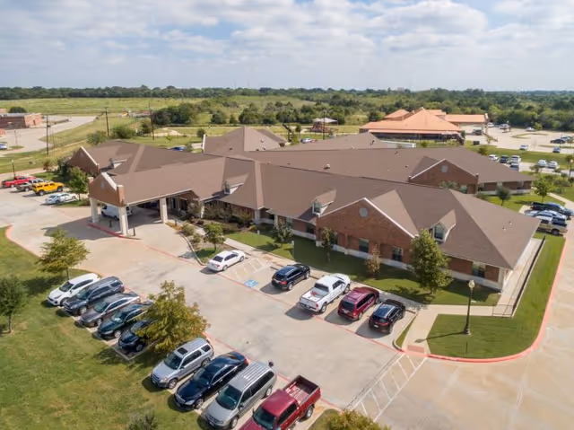 Aerial view of a single-story brick healthcare and rehabilitation facility with a large parking lot in front. Several cars are parked in the lot, and the building is surrounded by green lawns and trees. The sky is partly cloudy.