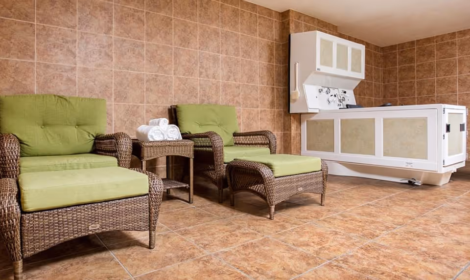 Tiled wellness room with wicker lounge chairs with green cushions, a side table with rolled towels, and a white hydrotherapy tub.
